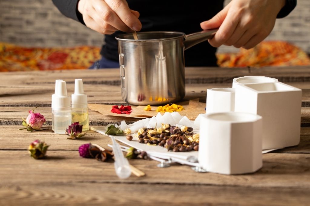 Person making candle, with fragrance oil, flower petals, and containers on a wooden table