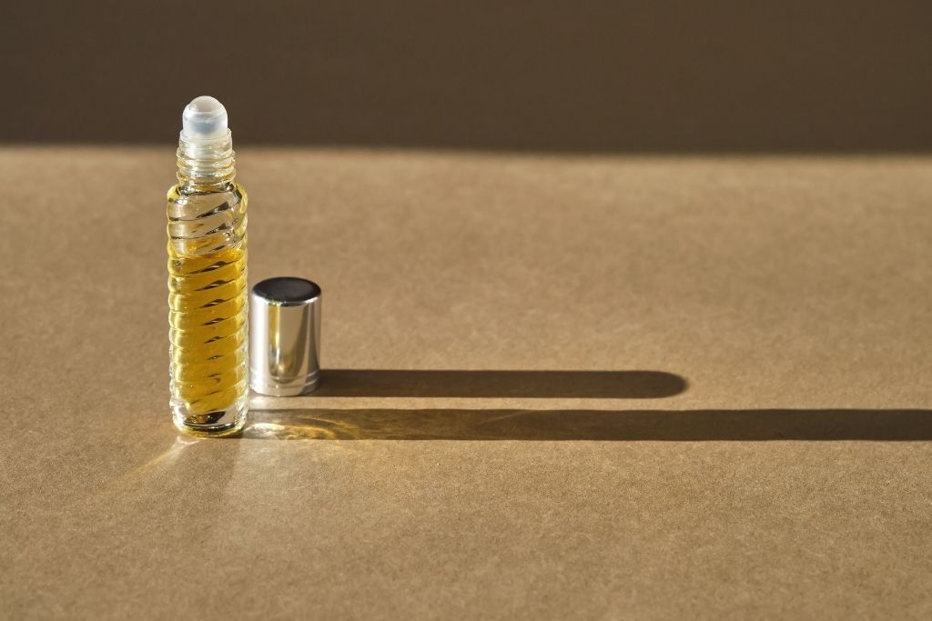glass roll-on perfume bottle filled with yellow-tinted oil, casting a long shadow on a brown background. The silver cap is sitting next to the bottle.