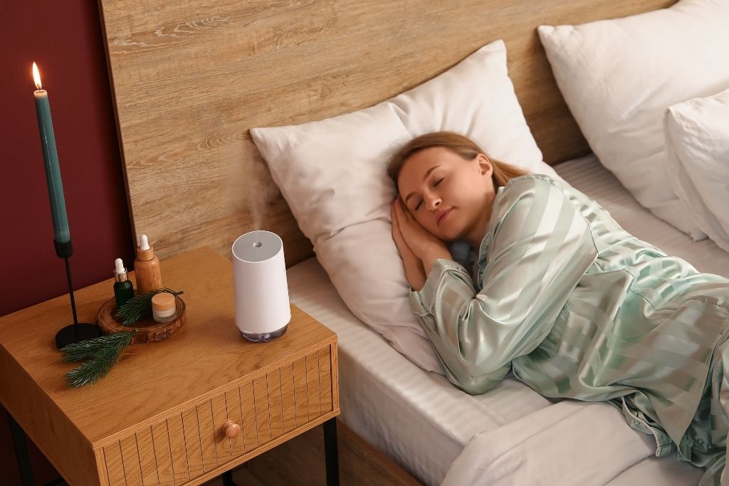 A woman in pajamas sleeping peacefully in bed with a white essential oil diffuser misting on her nightstand alongside a lit candle and small bottles.